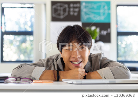Smiling asian teenage boy with braces leaning on desk in school classroom, feeling relaxed Smiling asian teenage boy with braces leaning on desk in school classroom, feeling relaxed 120369368