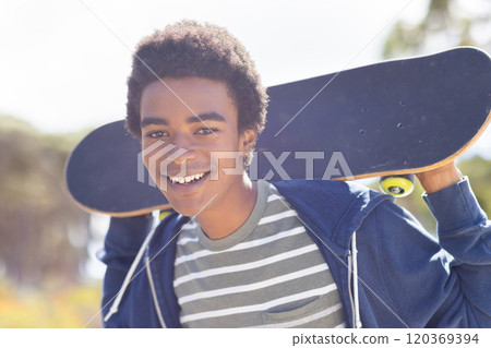 Smiling african american teenage boy holding skateboard, enjoying outdoor hangout on sunny day 120369394