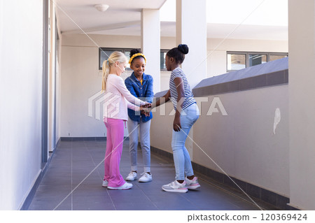 In school, three multiracial girls standing together in hallway, holding hands 120369424