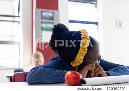In school, african american girl resting head on desk with apple and notebook nearby 120369501