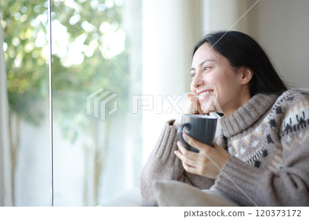 Asian woman drinking coffee in winter looking through a wiindow 120373172