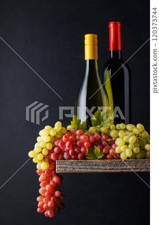 Bottles of red and white wine paired with grapes, set against a dark background 120373744