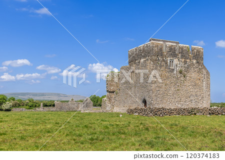 The tower house at Kilmacduagh Abbey in County Galway, Ireland, stands prominently in a green field under a clear blue sky. The medieval stone structure, part of the abbey historic ruins The tower house at Kilmacduagh Abbey in County Galway, Ireland, stands prominently in a green field under a clear blue sky. The medieval stone structure, part of the abbey historic ruins 120374183
