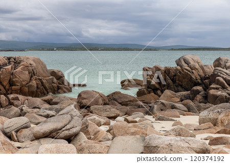 Massive boulders dominate the shoreline of Gurteen Beach, Ireland, leading to tranquil turquoise waters. The overcast sky and distant, rolling hills contribute to the rugged and serene atmosphere 120374192