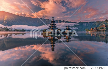 A serene Balinese temple reflecting in calm lake waters under a breathtaking sunrise. Misty mountains and soft clouds enhance the spiritual and tranquil atmosphere 120374925