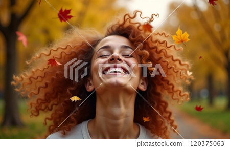 A joyful moment captured as a woman with vibrant curly hair laughs with pure happiness, surrounded by falling autumn maple leaves in a golden-hued park setting 120375063