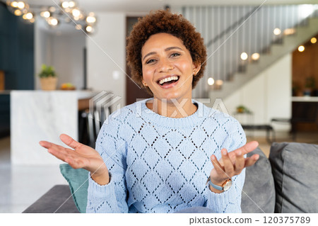 Smiling woman in blue sweater gesturing while sitting on couch at home, on video call 120375789