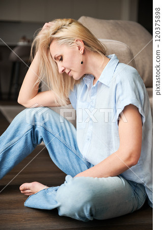 Side view of pretty woman looking down. Female with blonde hair relaxing next to couch. Beautiful woman sitting on wooden floor. Side view of pretty woman looking down. Female with blonde hair relaxing next to couch. Beautiful woman sitting on wooden floor. 120375898