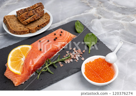 Fillet of raw salmon fish with arugula, basil and lemon on the cutting board with red caviar. 120376274