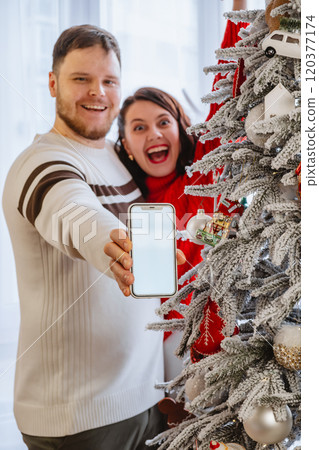 man with woman holding phone with empty screen standing near christmas tree 120377174