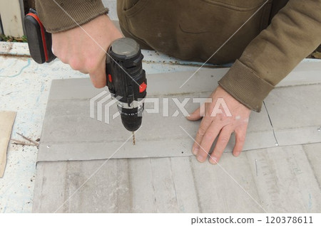 A construction worker fastens gray metal sheets together that are lying on a blue rusty table, using a screwdriver and self-tapping screws A construction worker fastens gray metal sheets together that are lying on a blue rusty table, using a screwdriver and self-tapping screws 120378611