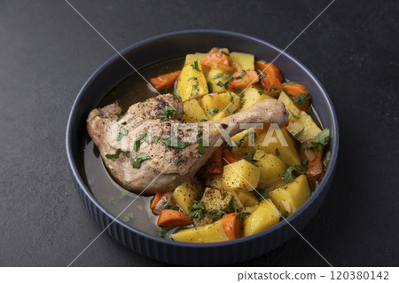 Duck leg with broth, stewed with potatoes and carrots, sprinkled with parsley and sumac. Simple home hearty cooking. Deep gray plate on a black background. Selective focus, close-up. 120380142