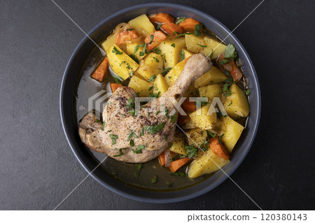 Duck leg with broth, stewed with potatoes and carrots, sprinkled with parsley and sumac. Simple home hearty cooking. Deep gray plate on a black background. Selective focus, close-up. Duck leg with broth, stewed with potatoes and carrots, sprinkled with parsley and sumac. Simple home hearty cooking. Deep gray plate on a black background. Selective focus, close-up. 120380143