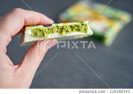 Piece of trend Dubai white chocolate with pistachio paste and kataifi dough in woman hand. Confectionery handmade sweets at home in the kitchen. Piece of trend Dubai white chocolate with pistachio paste and kataifi dough in woman hand. Confectionery handmade sweets at home in the kitchen. 120380343