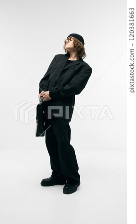 Full-length portrait of confident young guy in beanie hat, black costume, glasses and leather bag, posing against white studio background 120381663