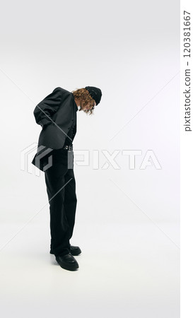 Side view full-length image of young man wearing total black outfit, blazer, pants and beanie hat, posing against white studio background 120381667
