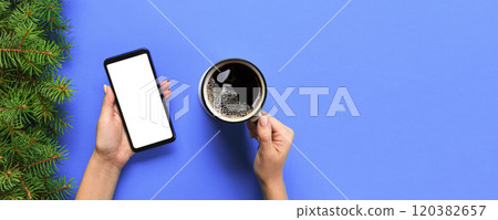 Female hands holding black mobile phone with blank white screen and mug of coffee. Mockup image with copy space. Top view on purple background, flat lay 120382657