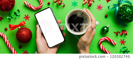 Above view of female hand holding smart phone with hot cup of coffee. Christmas decoration on colored background 120382748