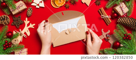 Top view of woman holding an envelope on red background made of holiday decorations. Christmas time concept Top view of woman holding an envelope on red background made of holiday decorations. Christmas time concept 120382850