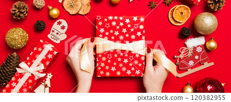 Top view of female hands holding a Christmas present on festive red background. Holiday decorations. New Year holiday concept Top view of female hands holding a Christmas present on festive red background. Holiday decorations. New Year holiday concept 120382955