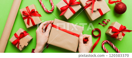 Woman is packing surprise gift for Christmas holidays, sitting at workplace with craft tools, top view. Woman wrapping Christmas gift at colored table 120383370
