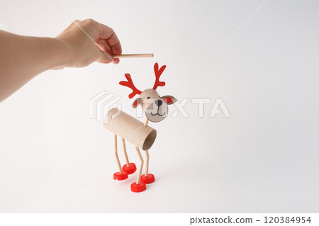 Hand holding DIY reindeer puppet made from cardboard tubes, plastic caps, and paper details. Christmas craft and holiday decoration concept, easy activity to do, paper roll craft for kid, 120384954