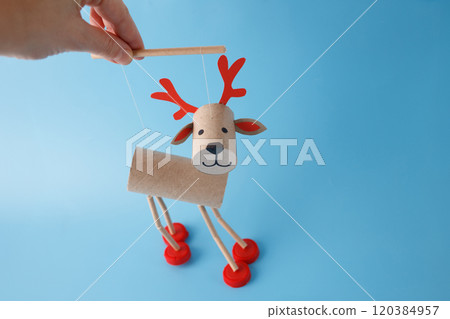 Hand holding DIY reindeer puppet made of cardboard tubes, plastic caps, and paper. on a blue background. Christmas craft and holiday decoration concept. marionette from recycled materials, Hand holding DIY reindeer puppet made of cardboard tubes, plastic caps, and paper. on a blue background. Christmas craft and holiday decoration concept. marionette from recycled materials, 120384957