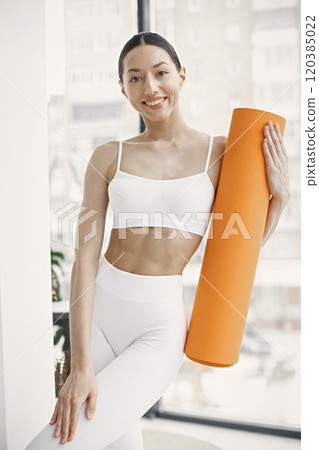 Slim and beautiful woman wearing white leggins and top and holding rolling yoga mat. Young girl with fit body posing in studio with big windows. Sport, fitness, diet, nutrition and health care concept Slim and beautiful woman wearing white leggins and top and holding rolling yoga mat. Young girl with fit body posing in studio with big windows. Sport, fitness, diet, nutrition and health care concept 120385022