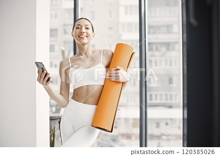 Slim and beautiful woman wearing white leggins and top and holding rolling yoga mat. Young girl with fit body posing in studio with big windows and using a phone. Sport, fitness, diet, nutrition and 120385026