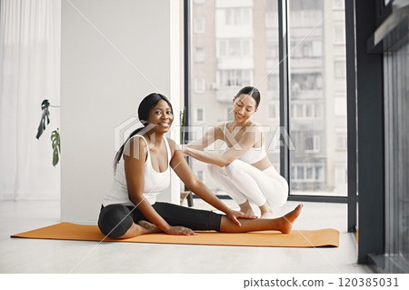 Two women working out on yoga mat in bright studio with big windows. Caucasian woman wearing white leggins and top and black woman white t-shirt and black leggins. Woman and her coach in gym. Two women working out on yoga mat in bright studio with big windows. Caucasian woman wearing white leggins and top and black woman white t-shirt and black leggins. Woman and her coach in gym. 120385031