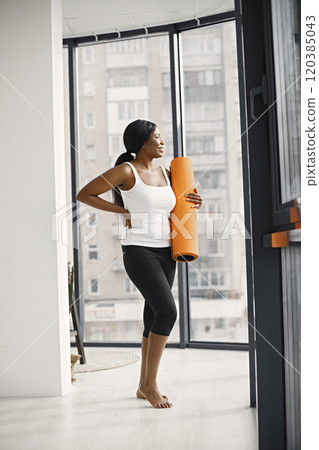 Black woman wearing black leggins and white top and holding rolling yoga mat. Young girl with fit body posing in studio with big windows. Sport, fitness, diet, nutrition and health care concept. Black woman wearing black leggins and white top and holding rolling yoga mat. Young girl with fit body posing in studio with big windows. Sport, fitness, diet, nutrition and health care concept. 120385043
