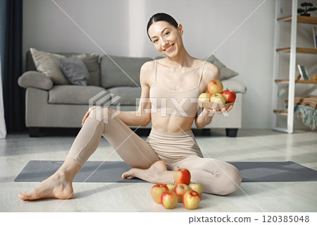 Slim and beautiful woman wearing beige leggins and top and sitting on yoga mat. Young girl with fit body posing at home with fresh apples. Sport, fitness, diet, nutrition and health care concept. Slim and beautiful woman wearing beige leggins and top and sitting on yoga mat. Young girl with fit body posing at home with fresh apples. Sport, fitness, diet, nutrition and health care concept. 120385048