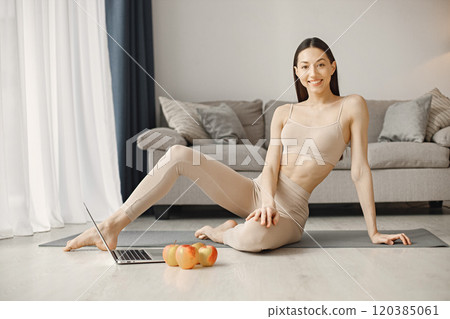 Slim and beautiful woman wearing beige leggins and top and sitting on yoga mat. Young girl with fit body posing at home with fresh apples. Sport, fitness, diet, nutrition and health care concept. 120385061