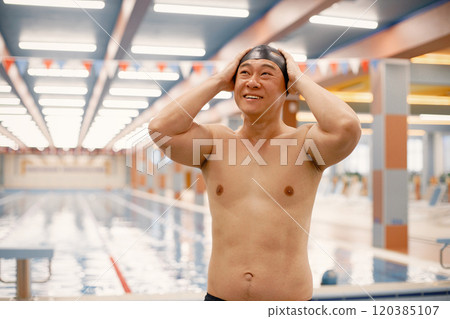 Man wearing special hat for swimming. Asian man standing in indoor swimming pool. Professionale male swimmer is going to swim. 120385107