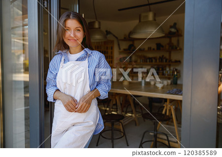 A cheerful woman wearing an apron stands in a cozy workshop space filled with creativity and warmth 120385589