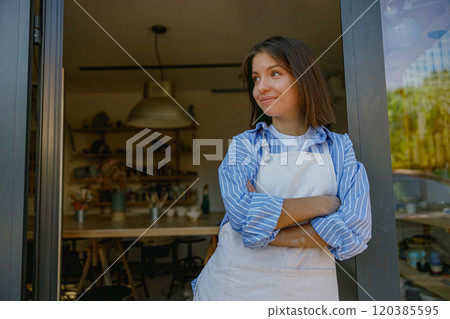 A serene woman in an apron stands at her creative studios entrance, ready for a productive day 120385595