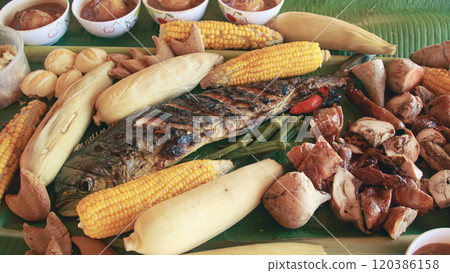 A close-up view of a traditional Filipino sharing meal called boodle fight with grilled fish, chicken corn, and assorted vegetables served on a banana leaf 120386158