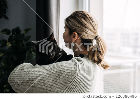 Woman holding black cat close by the window, symbolizing pet love and companionship. Calm, nurturing moment between owner and furry friend. 120386490