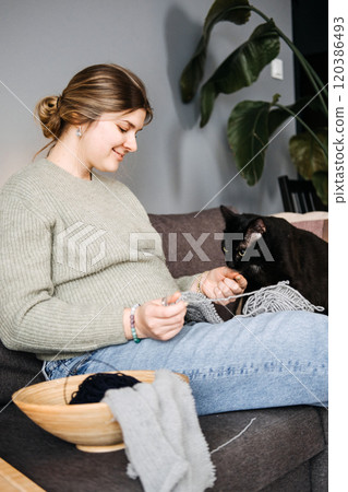 Pregnant woman knitting with black cat beside, preparing handmade baby items. Cozy nesting, relaxation, and mindful pregnancy. 120386493
