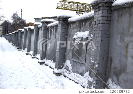 A long grey concrete fence by a snow way in front of an industrial wall of a factory in Ukraine in city Sumy A long grey concrete fence by a snow way in front of an industrial wall of a factory in Ukraine in city Sumy 120389782