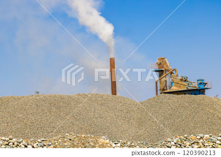 white steam rising up from pipe of small asphalt factory at day time with piles of gravel in a foreground 120390213