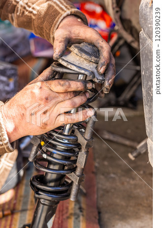 The calloused hands of a mechanic assembling compressed coil spring during DIY car repairs. The calloused hands of a mechanic assembling compressed coil spring during DIY car repairs. 120390239