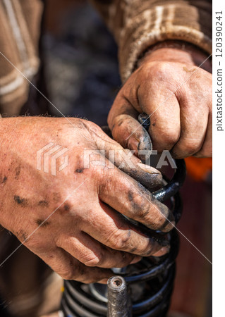 The calloused hands of a mechanic assembling compressed coil spring during DIY car repairs. The calloused hands of a mechanic assembling compressed coil spring during DIY car repairs. 120390241