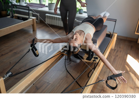 A Pilates Instructor helps a client with stretching exercises on reformer bed to improve flexibility and wellbeing A Pilates Instructor helps a client with stretching exercises on reformer bed to improve flexibility and wellbeing 120390380