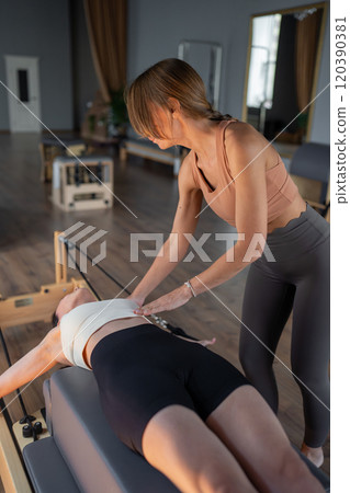 A Pilates Instructor helps a client with stretching exercises on reformer bed to improve flexibility and wellbeing  120390381
