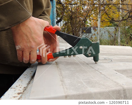 male hands gripping riveting tool handle with force, fixing aluminum riveting into metal structure, fastening layers of thin iron with hand riveter side view 120390711