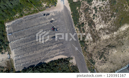 An aerial view of a mostly empty paved parking area near a sandy, grassy landscape, with a few bicycles and people gathered. Fenced bike racks line the parking area, surrounded by greenery. An aerial view of a mostly empty paved parking area near a sandy, grassy landscape, with a few bicycles and people gathered. Fenced bike racks line the parking area, surrounded by greenery. 120391167