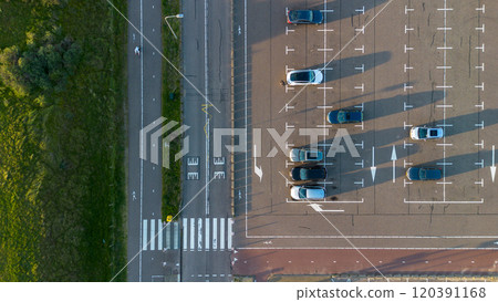 Aerial view of a parking lot with parked cars casting long shadows under soft sunlight. Adjacent to the lot, there is a green grassy area and a pedestrian walkway with a zebra crossing. Aerial view of a parking lot with parked cars casting long shadows under soft sunlight. Adjacent to the lot, there is a green grassy area and a pedestrian walkway with a zebra crossing. 120391168