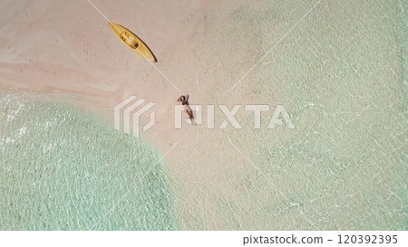 Aerial view of a serene tropical paradise featuring a woman enjoying the sun on a secluded pink sand beach with a kayak nearby 120392395