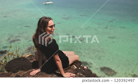 Young tourist is enjoying the view of the ocean from a cliff on a sunny day. The water is a beautiful turquoise color and the sun is shining brightly Young tourist is enjoying the view of the ocean from a cliff on a sunny day. The water is a beautiful turquoise color and the sun is shining brightly 120392497